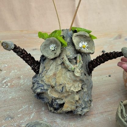 Töpfern für Kinder Eine kreative Figur aus Holz, mit Zweigen als Armen und Blumen als Augen.